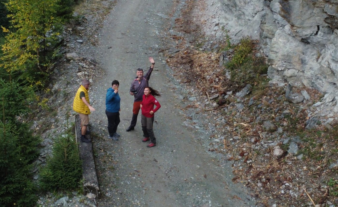 Drone view of Alex and Elisa with Radu and Violeta waving on a forest road below a rock face