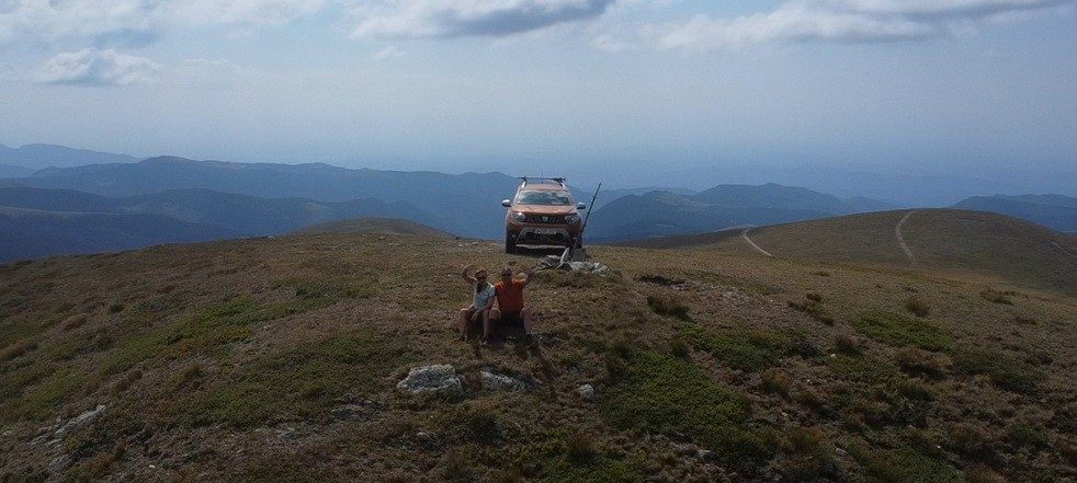 Two people sitting on a summit, with the car parked on the ridge behind them